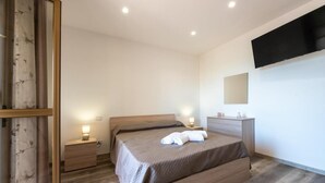 A modern bedroom with a wooden bed, bedside tables with lamps, a flat-screen TV mounted on the wall, and a large mirror.