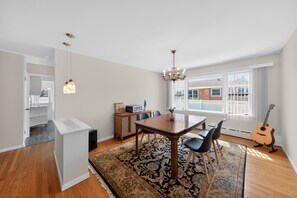 Un espace repas comprenant une table, des chaises, un lustre, une guitare et une vue dégagée sur l’extérieur grâce à de grandes fenêtres.