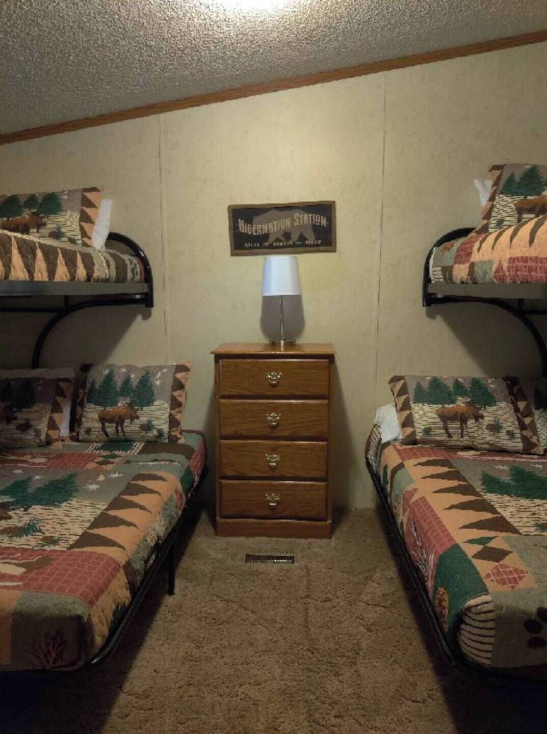 Una habitación con dos literas, cada una con una colcha estampada, un aparador de madera con cajones y una lámpara fijada a la pared.