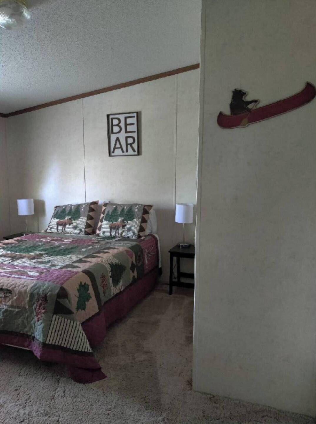 Un dormitorio con una cama, una colcha, un letrero en la pared, una mesita de noche y un adorno de pared.