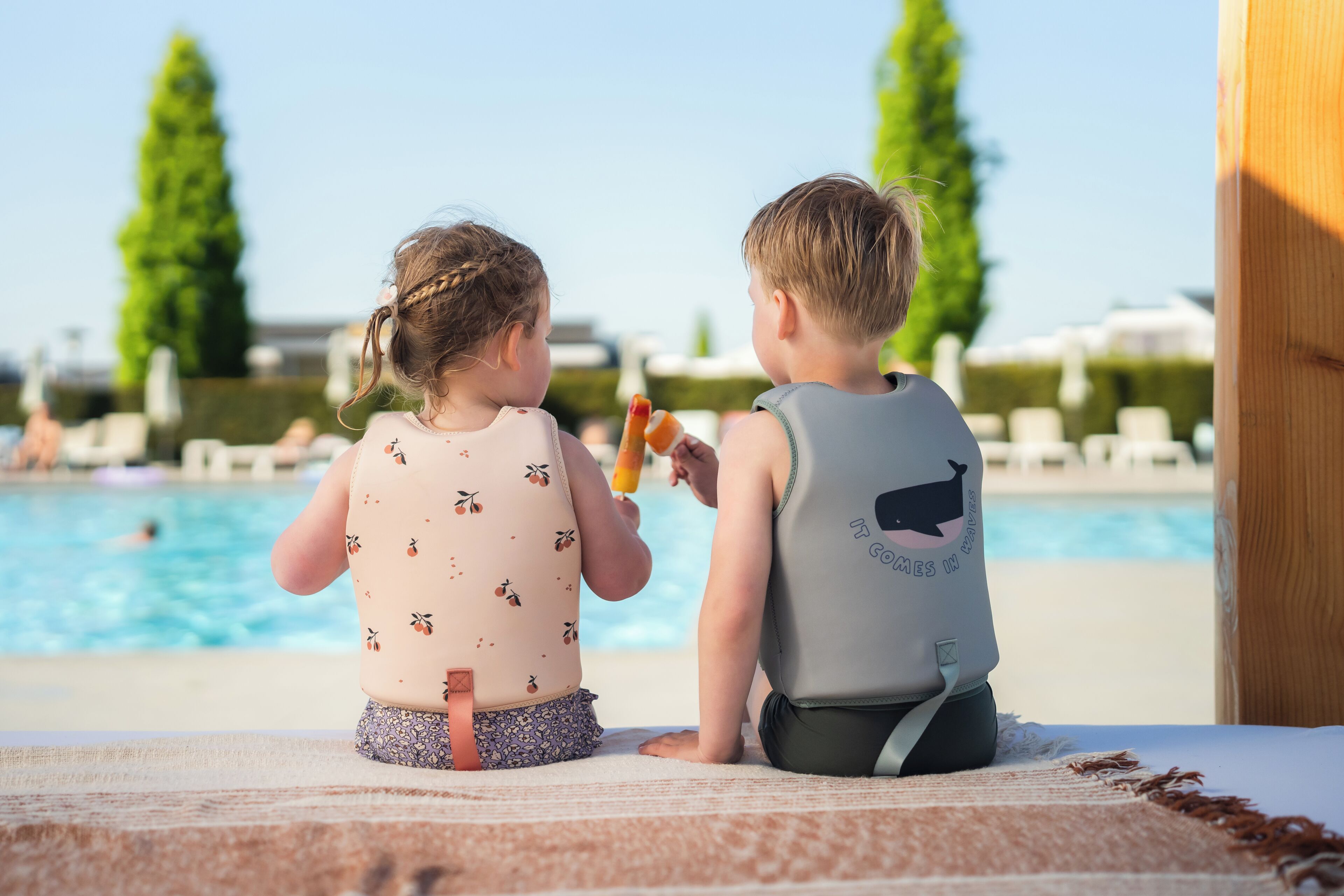 Twee kinderen zitten bij een zwembad, met een helder uitzicht op het zwembad en ligbedden op de achtergrond.
