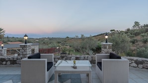 An outdoor seating area with wicker furniture and stone walls, overlooking a hilly landscape with vegetation and distant buildings. - Amara Haven (Aigialeia)