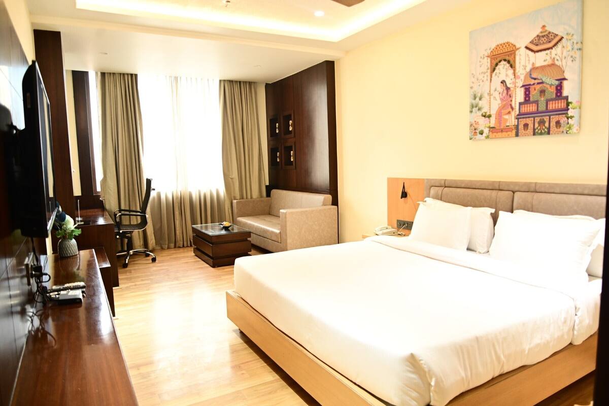 A hotel room with a large bed, a desk with a computer, a chair, a sofa, and a painting on the wall.