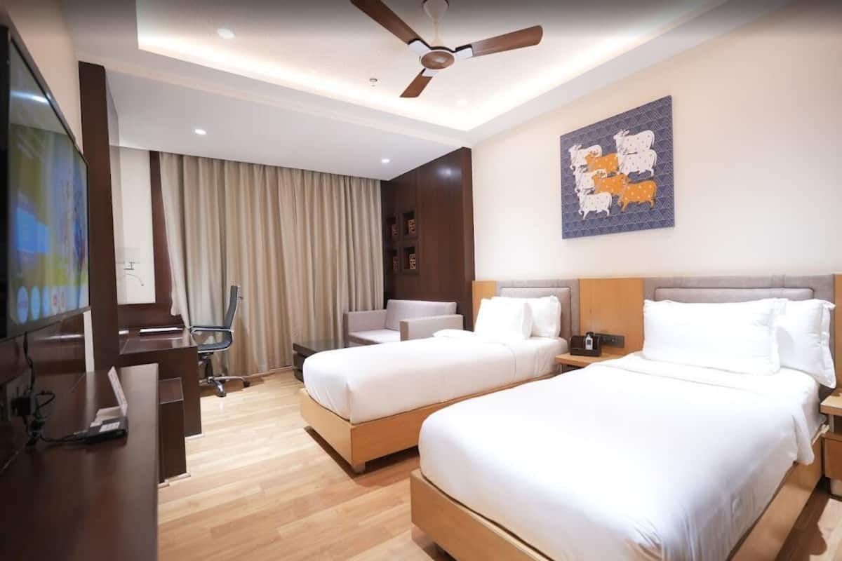 A hotel room with two beds, a desk, a chair, a TV, and a ceiling fan.