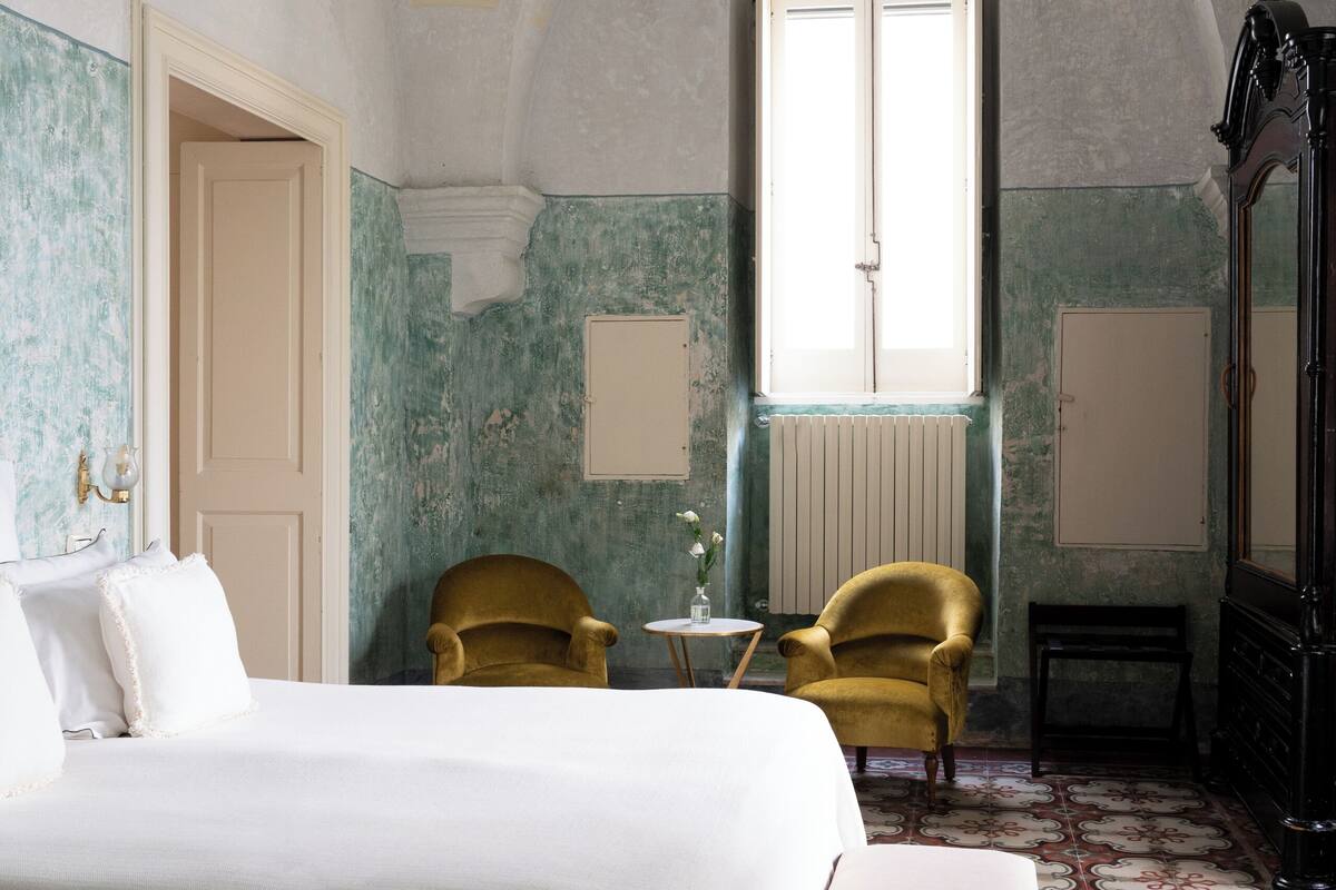 A bedroom with a bed, two chairs, a bench, a dresser, and a large window.