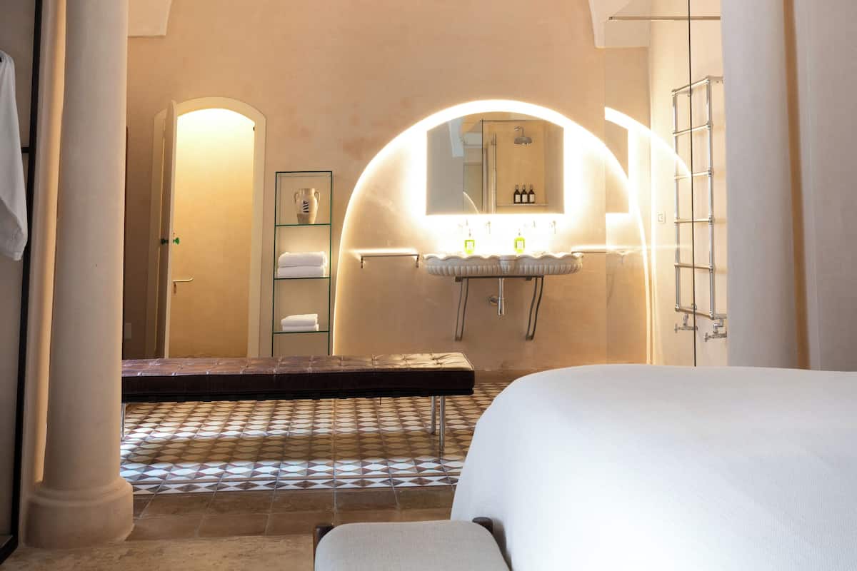 A hotel room with a large bed, a sitting area, a bathroom with a mirror, and a tiled floor.