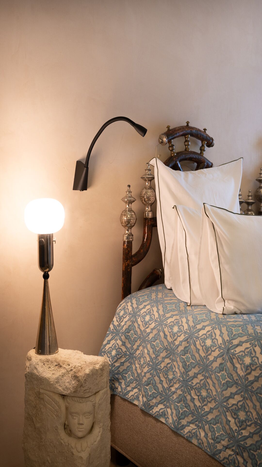 A bedroom with a patterned bedspread, a bedside lamp, a decorative wall-mounted object, and a stone sculpture.