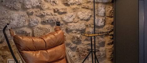 A leather armchair with a high backrest and a single black metal base, positioned against a textured stone wall.