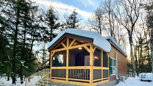 A wooden cabin with a covered porch and a stone foundation, surrounded by snow and trees.