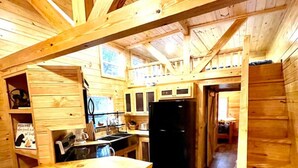 A wooden cabin interior with a kitchen area, wooden walls, and a loft bedroom.