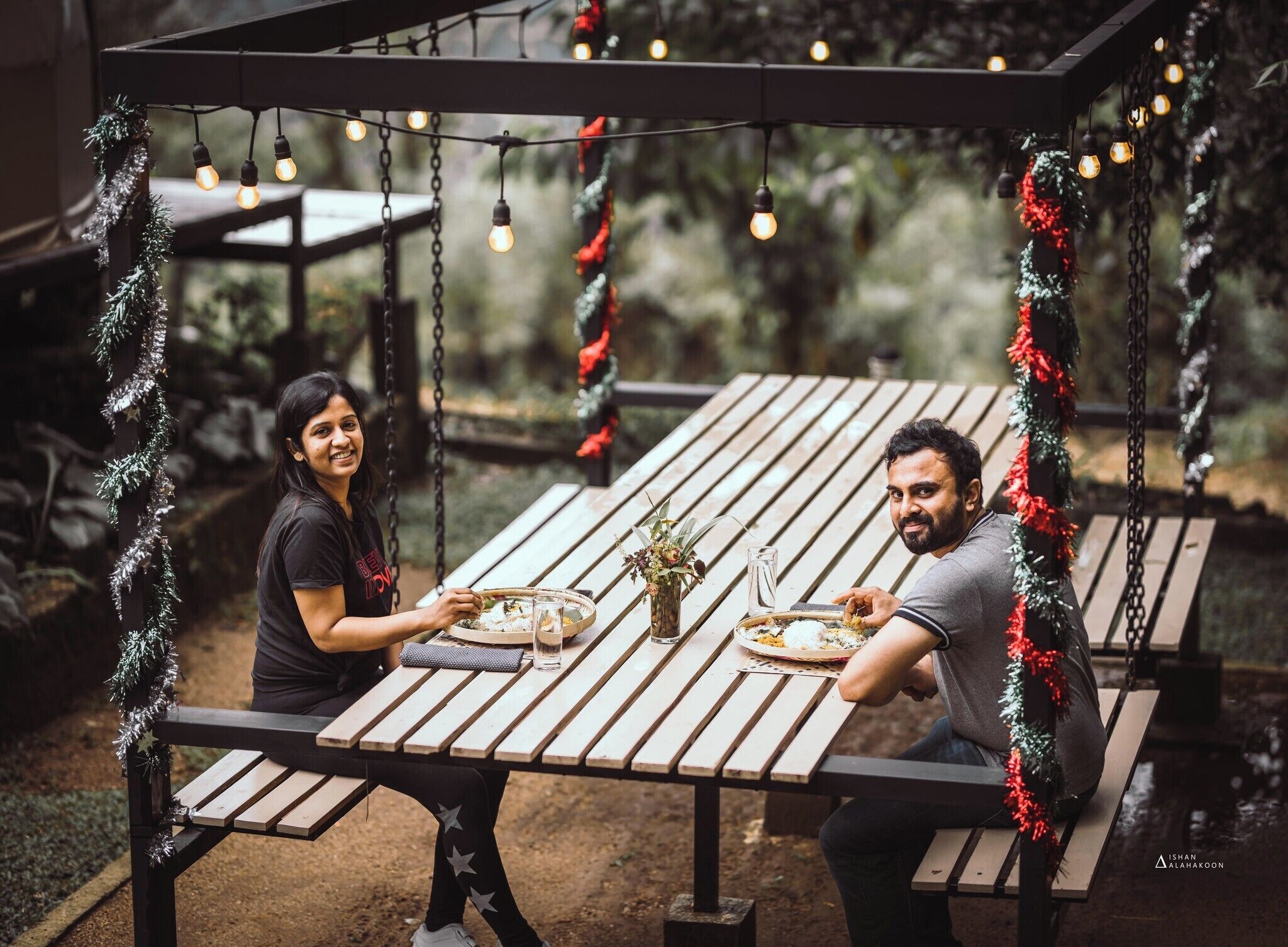 Dos personas cenando en una mesa de madera al aire libre con luces colgantes.