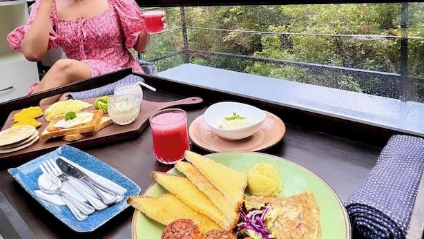 A dining table with food and a person in a pink top, overlooking a mountainous landscape. - Glamping by Offtrek (Medamahanuwara)