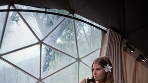 A person sitting on a bed inside a tent, holding a mug, with a tray of food and drinks in front. - Glamping by Offtrek (Medamahanuwara)