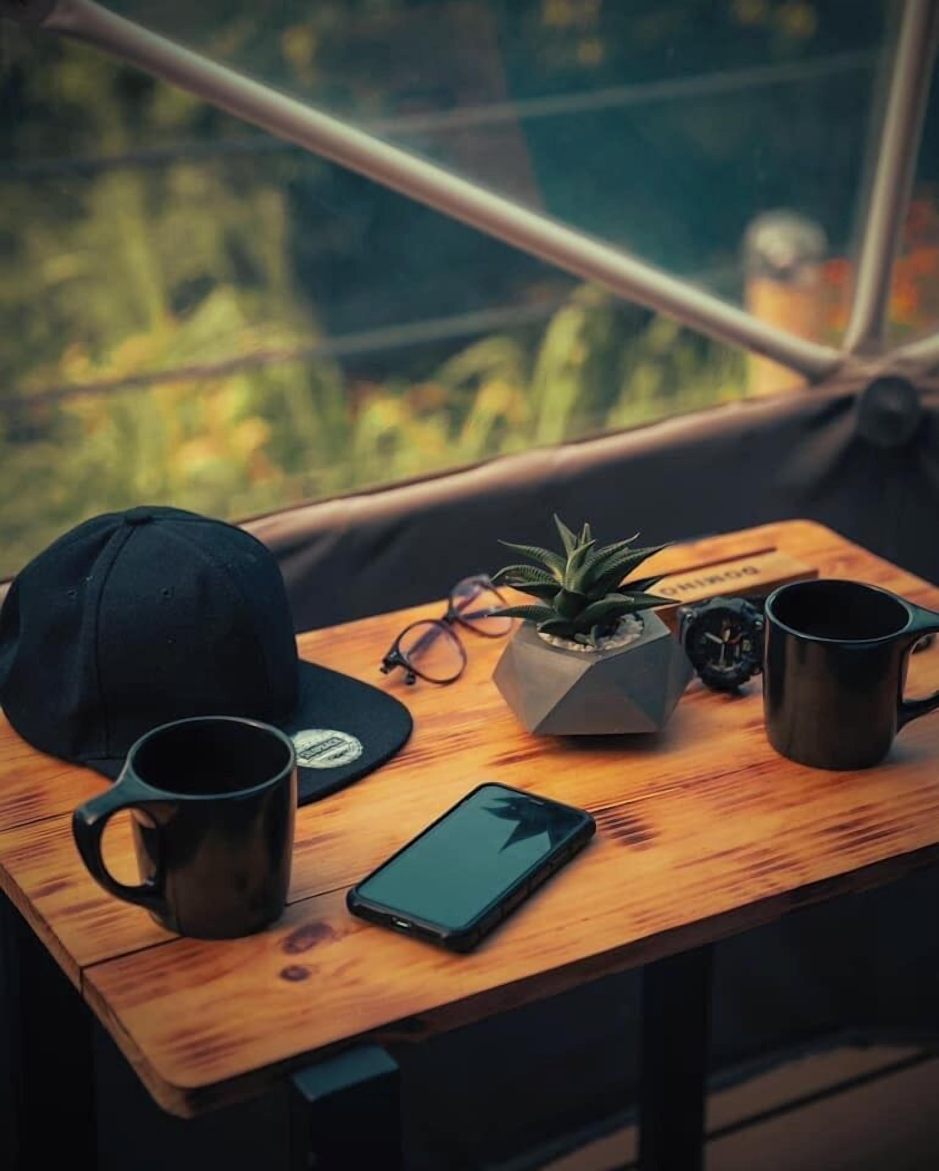 A wooden table with a smartphone, two black mugs, a black cap, a pair of glasses, and a small potted plant.