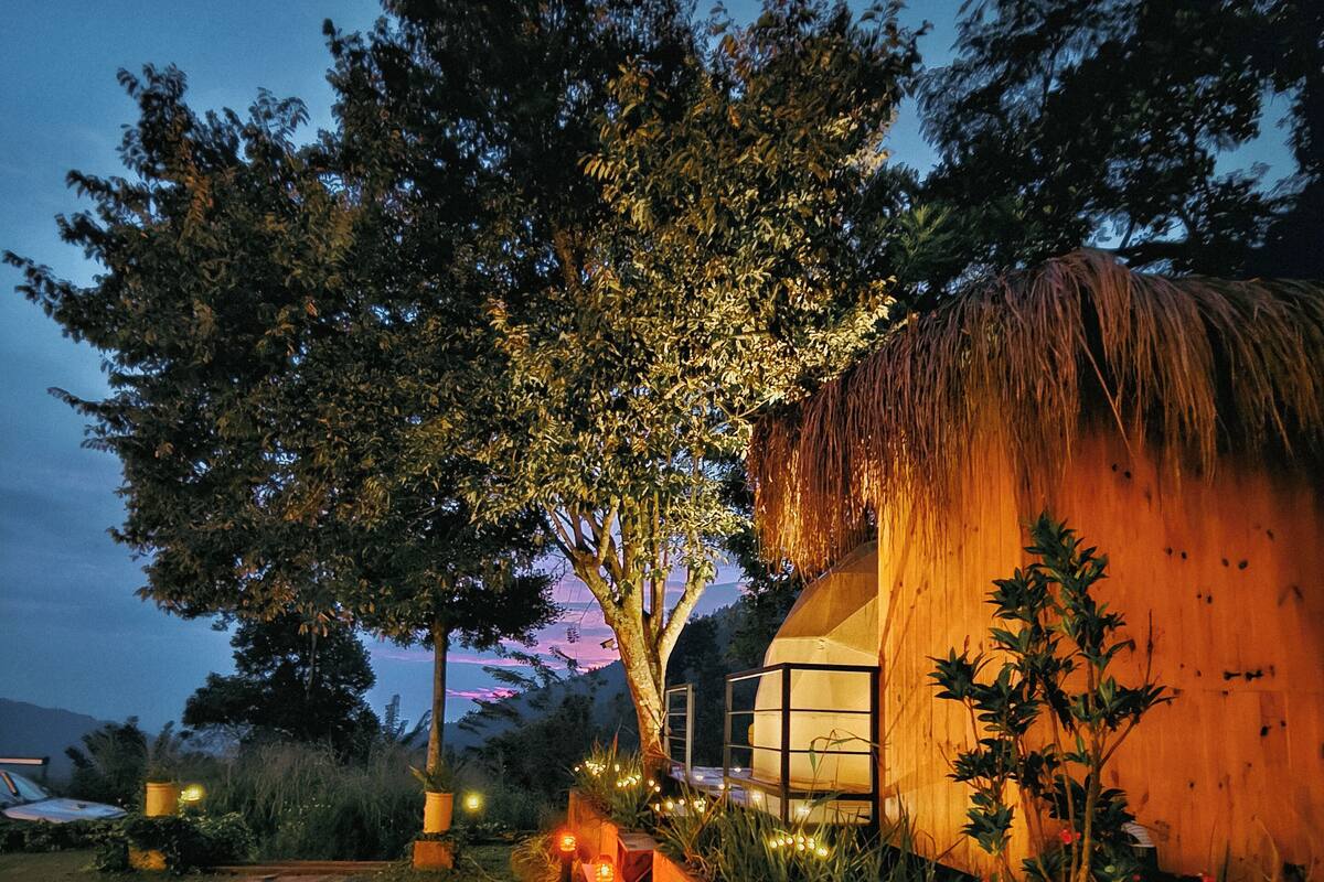 A thatched-roof structure with a glass extension, surrounded by a garden path and illuminated by string lights.
