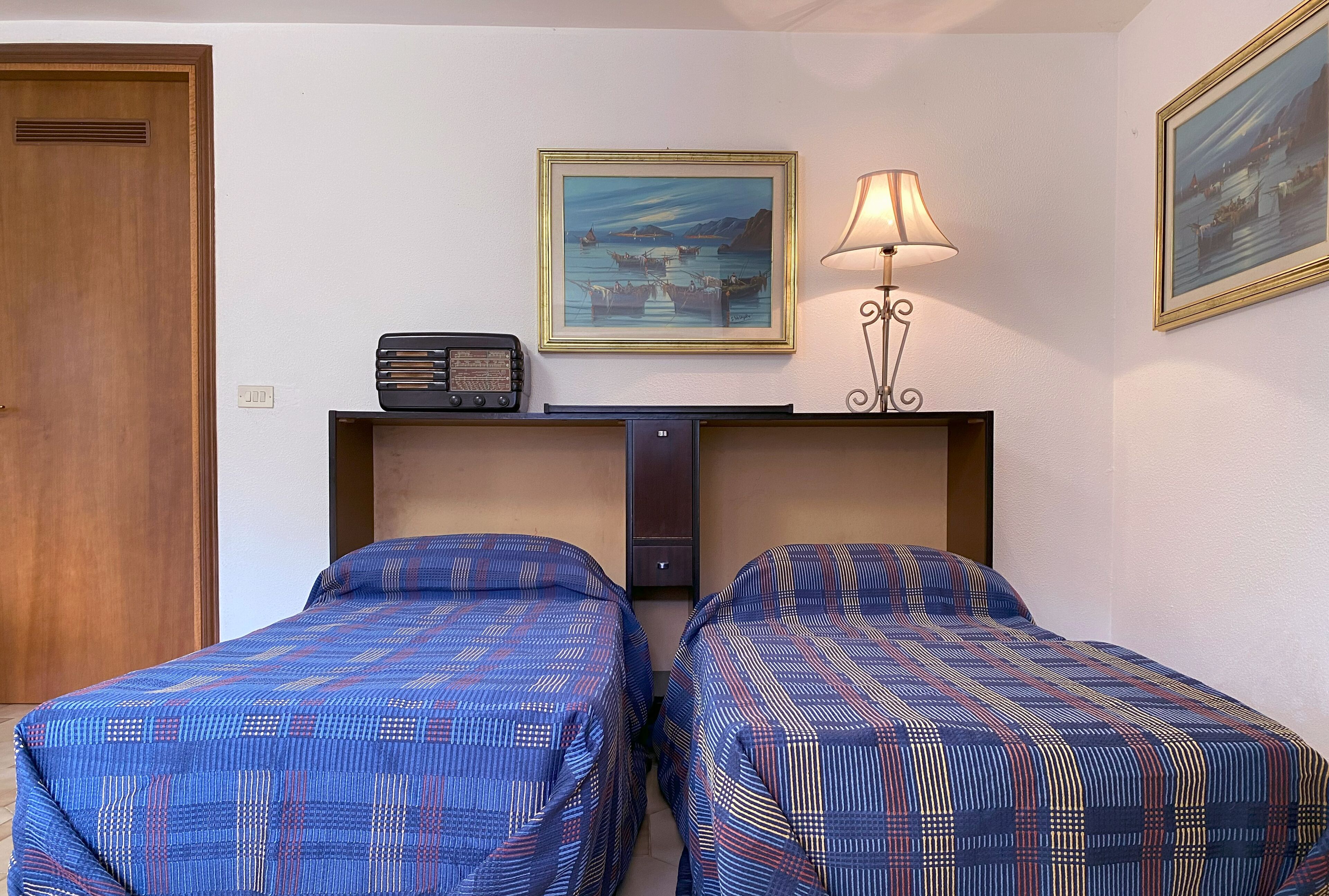 A room with two beds, a radio, a lamp, and two paintings on the wall.