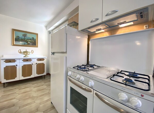 A compact kitchen with a gas stove, oven, and a white refrigerator. There is a wooden cabinet with a decorative item on top and a painting on the wall. - PERSEVERANTIA (Subiaco)