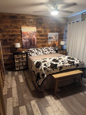 A bedroom with a wooden wall, a bed with a cow print blanket, two bedside lamps, and a ceiling fan. - Fayetteville, WV & Summersville, WV Proximity new built cabin w firepit & hottub (Nallen)