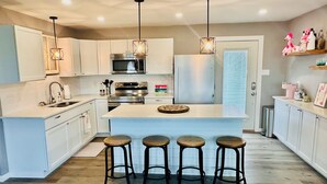 A modern kitchen with white cabinets, stainless steel appliances, a central island with stools, pendant lighting, and a view of the outdoors through a window.