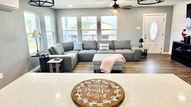 A modern living room with a sectional sofa, a ceiling fan, and a decorative wooden board with text.
