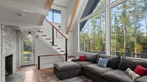 A modern living room with a fireplace, a staircase, and large windows offering a forest view. - Warren VT Chalet for Ski, Summer + Fall Fun, WIFI (Warren)