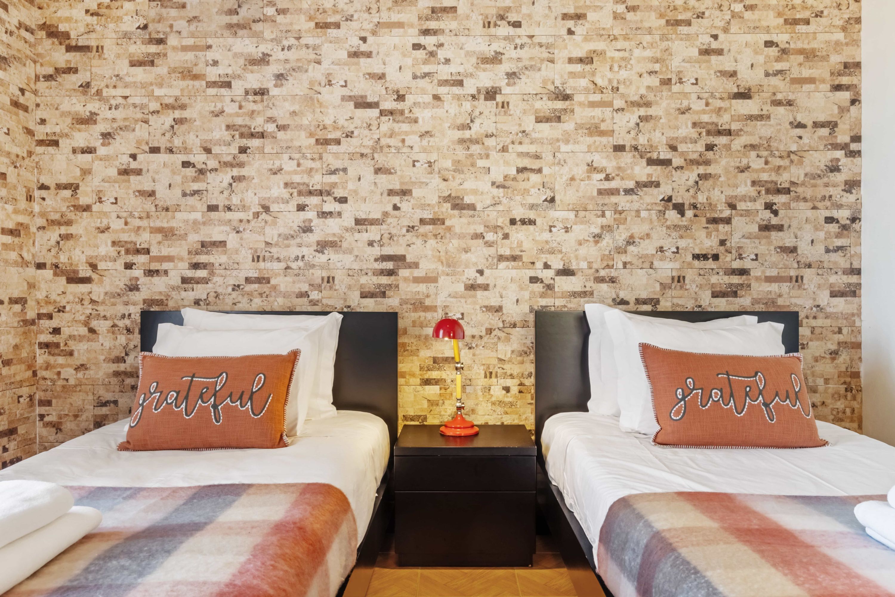 A hotel room with two beds, each with a pillow labeled "grateful," a nightstand with a lamp, and a brick-patterned wall.