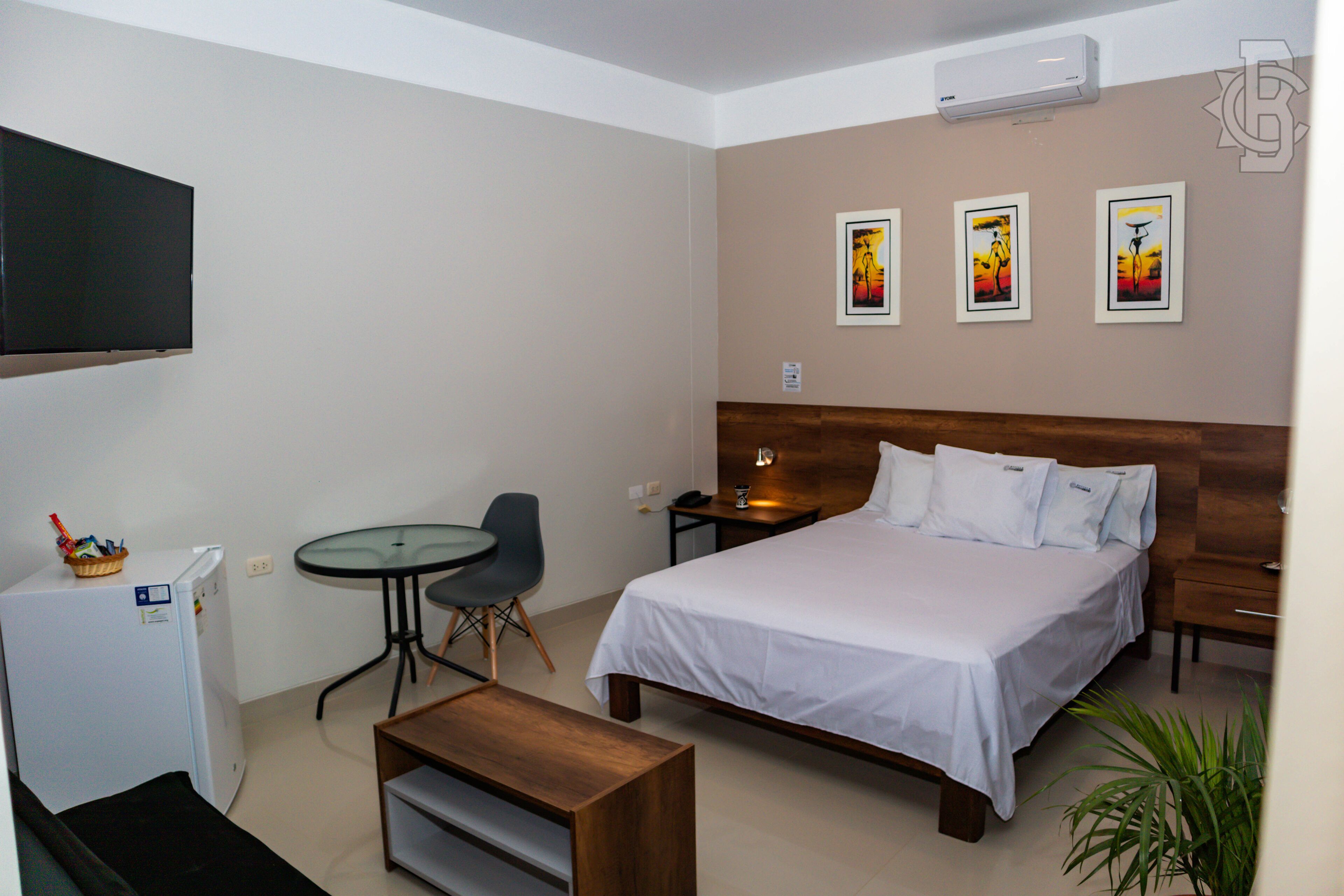 Una camera d'albergo con un letto, una scrivania, una sedia, un tavolino, un frigorifero e opere d'arte appese al muro.