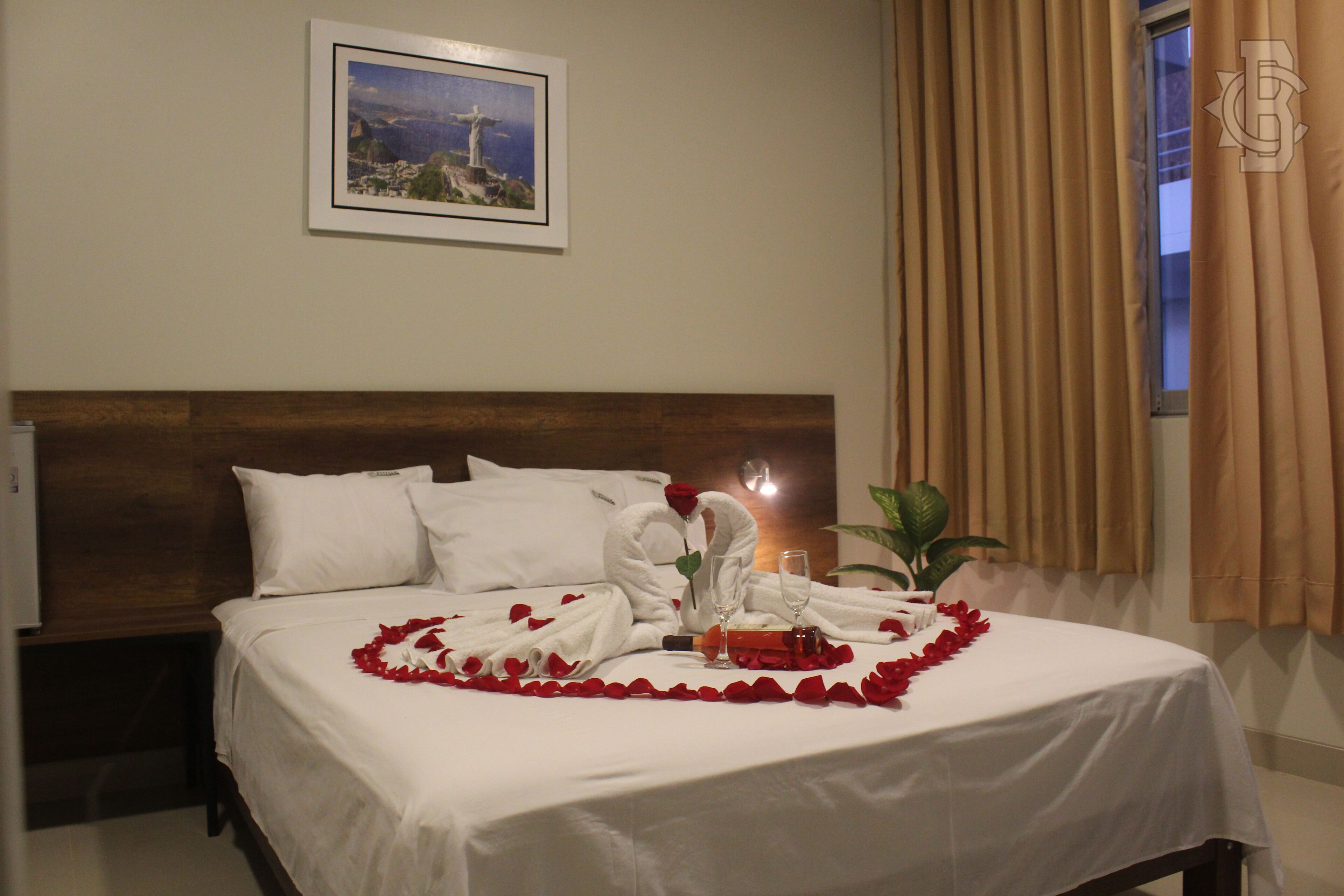 A neatly made bed with white linens, a heart-shaped arrangement of rose petals, and two glasses of champagne.