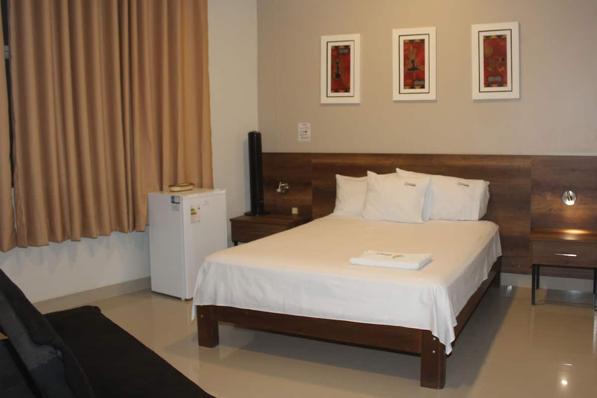 A hotel room with a large bed, a small bedside table, a nightstand, a refrigerator, and a window with curtains.