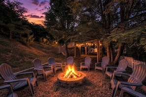 A fire pit surrounded by Adirondack chairs in a wooded outdoor setting.