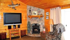 Ein gemütliches Wohnzimmer mit Kamin aus Stein, Holzwänden, einem Fernseher und einem gemusterten Sessel.