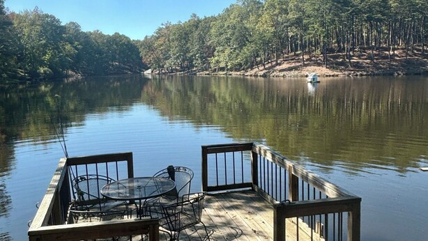 A wooden dock with a table and chairs, overlooking a calm lake surrounded by trees.