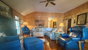 A cozy living room with wooden walls, a ceiling fan, a sectional sofa, a blue armchair, and a small round table with a lamp.