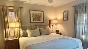 A bedroom with a bed, wooden floor, ceiling fan, nightstand, lamp, and a framed picture on the wall.