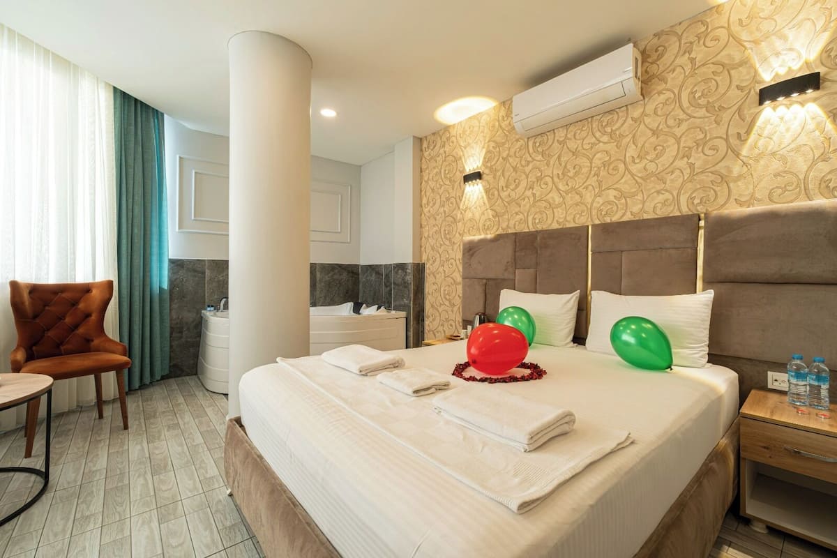 A hotel room with a large bed, a sitting area with a chair, a bathroom with a bathtub, and a wall-mounted air conditioner.