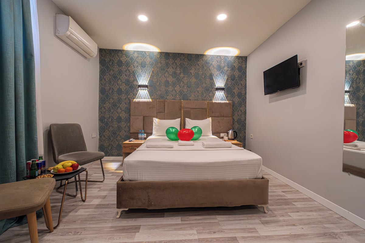 A hotel room with a bed, a chair, a table with fruit, an air conditioner, a wall-mounted TV, and patterned wallpaper.