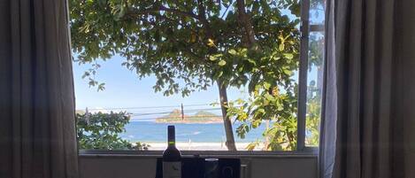 A room with a round table, two chairs, a bottle of wine, and a view of the sea through a window with curtains.