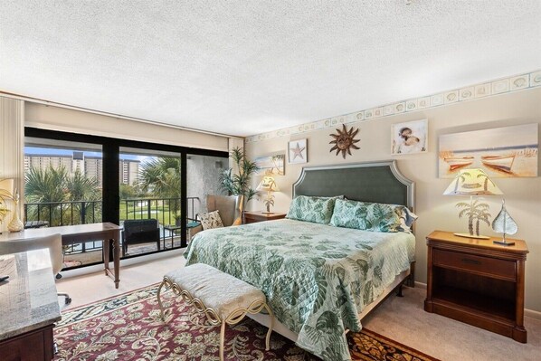 A bedroom with a bed, a desk, a chair, a balcony with a view, and a nightstand with a lamp.