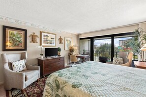 A hotel room with a large bed, a desk with a computer, a chair, a TV, and a balcony with a view of palm trees.