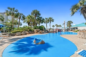 A swimming pool with a blue water feature, surrounded by palm trees and a building with a green roof.