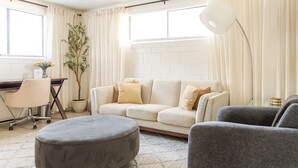 A living room with a sofa, ottoman, and armchair, a desk with a chair, a potted plant, and a window with curtains.