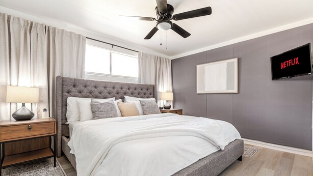 A bedroom with a tufted headboard, a bed with white bedding, a ceiling fan, a wall-mounted TV displaying 'NETFLIX', and a window with curtains.