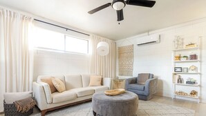 A living room with a sofa, ottoman, and armchair, a ceiling fan, and a white wall with shelves.
