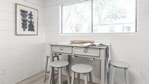 A white kitchen with a white brick wall, a window with blinds, a framed picture, and two stools.