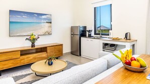 A modern kitchen with a refrigerator, stove, and sink. There is a dining table with a fruit bowl and a sofa in the living area. A large window provides a view of the beach.