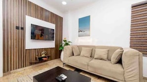 Een woonkamer met een beige bank, een flatscreen televisie bevestigd aan een houten wand, een salontafel, en een lijst met een afbeelding aan de muur.
