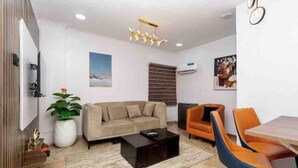 Een moderne woonkamer met een bank, salontafel en oranje stoelen. Er is een televisie, een plant en kunstwerken aan de muren.