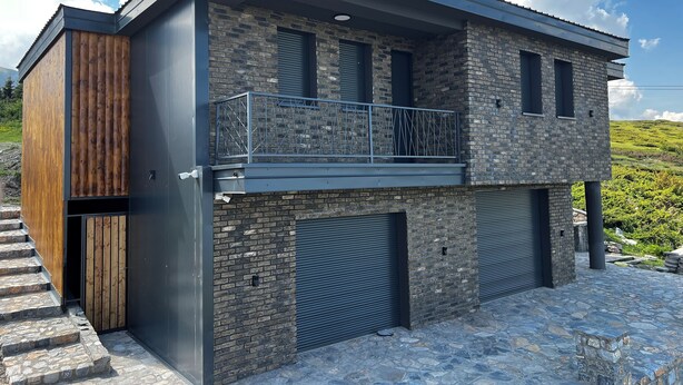 Une maison moderne à deux étages, dotée d’une façade en pierre, d’un balcon avec des rampes en métal et de deux portes de garage.