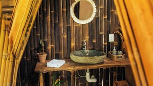 A bathroom with a bamboo wall, a round mirror, a sink, a potted plant, and a shelf with toiletries. - Bambuk Bio Chalet Uvita beach front (Uvita)