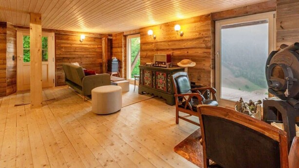 A cozy cabin interior with wooden walls and ceiling, a sofa, a dining table, and a large window offering a view of the outdoors. - Chalet in Rossiniere With Sauna and Mountain Views (Chateau-d'Oex)